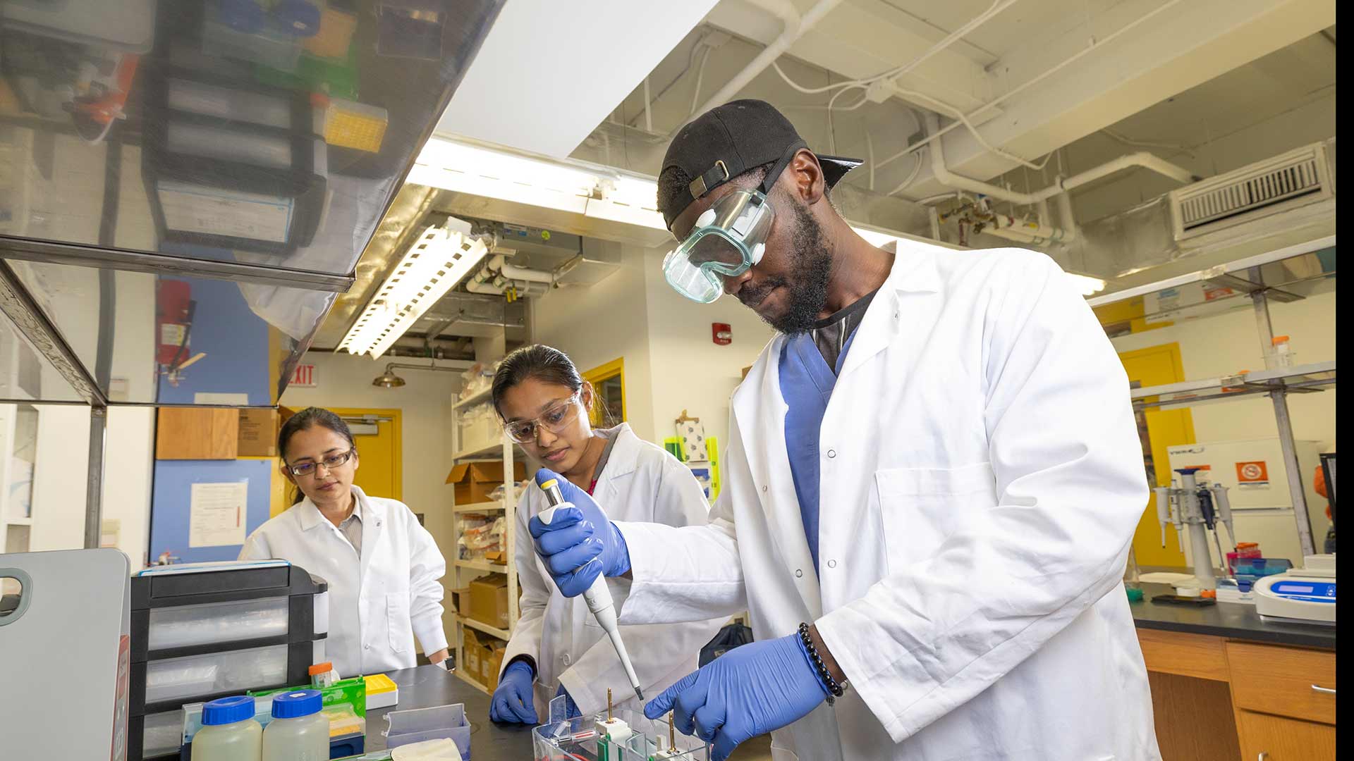 A chemistry graduate student works in a lab by mixing materials with a tool while another student and a professor observe.