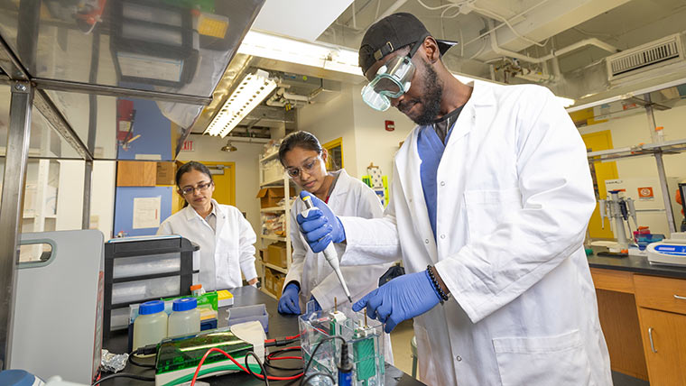 Three chemistry graduate students working in a lab. One is mixing materials with a tool while the other two observe.