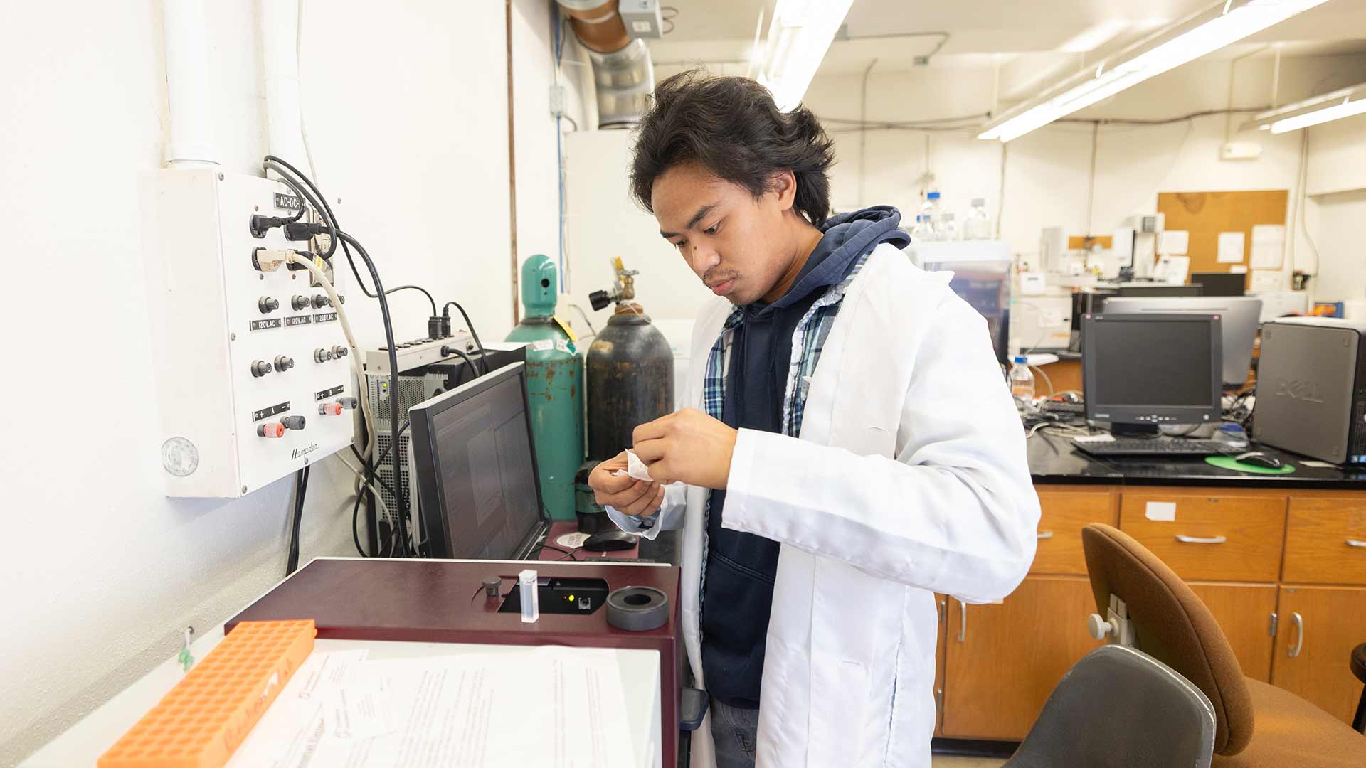 A student in a white lab coat handles chemical supplies in a lab.